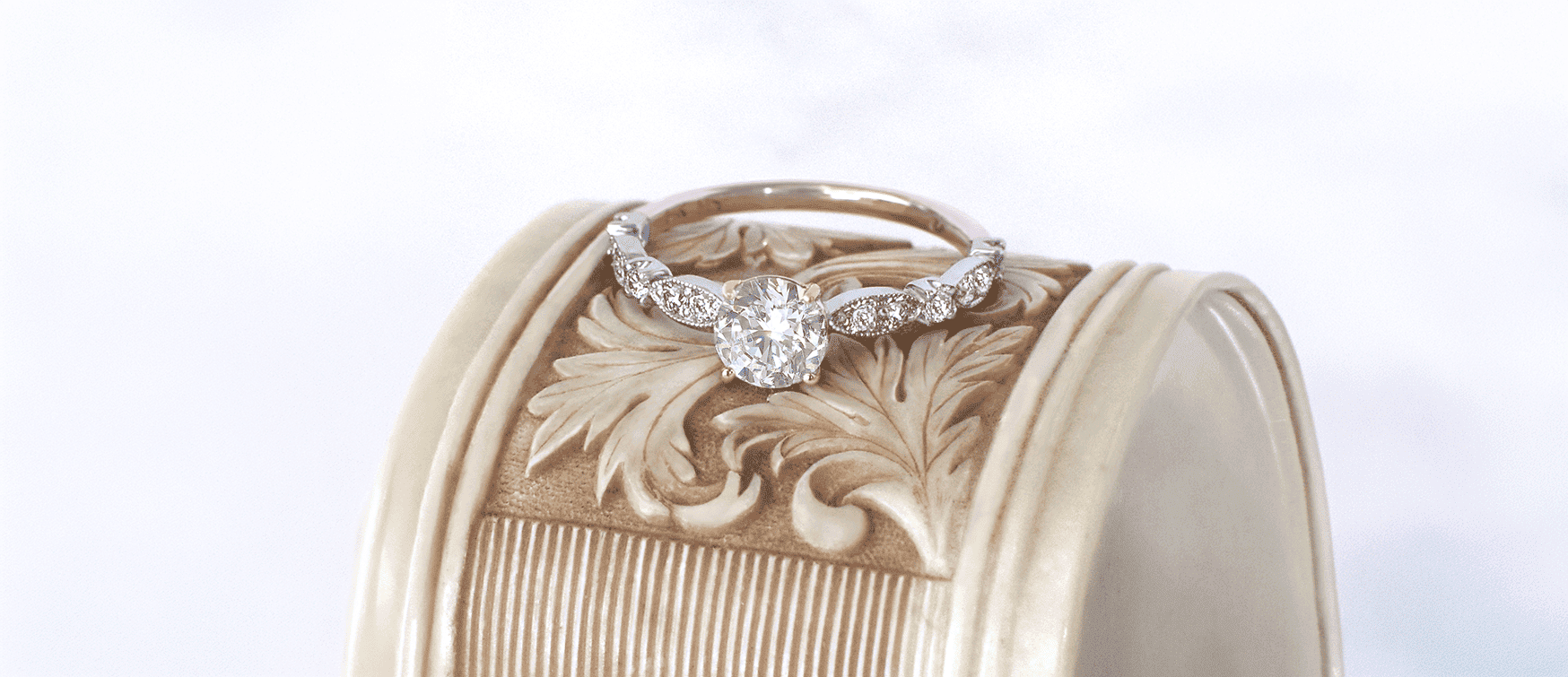 Vintage-inspired Round Brilliant Diamond Engagement Ring with Milgrain Shoulder Detailing Resting on an Ornate Carved Pedestal at Mark Solomon Jewellers.