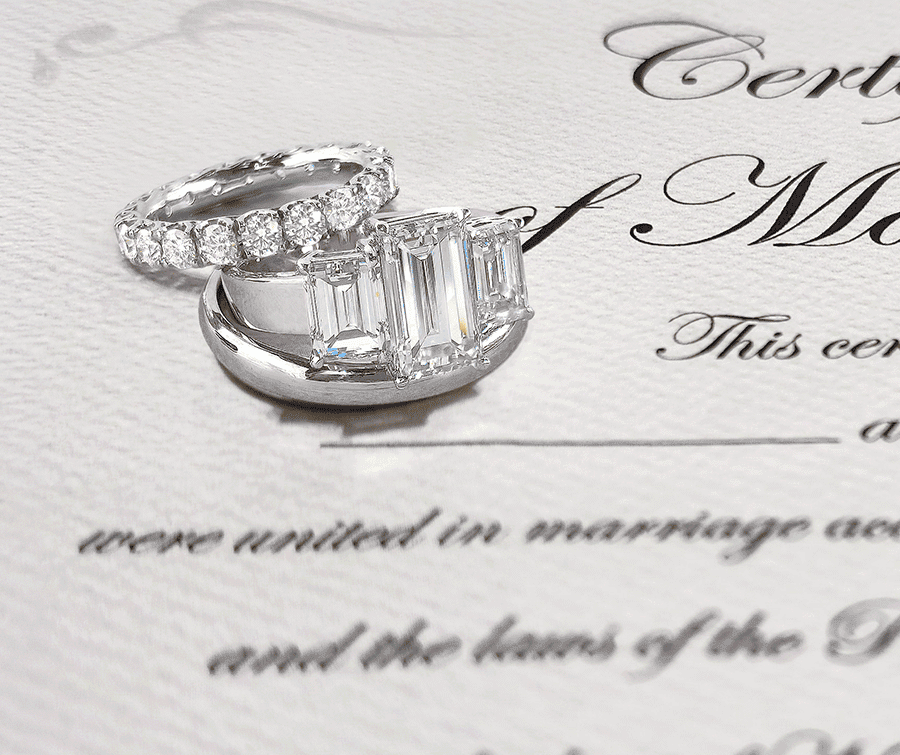 Three-stone emerald-cut diamond engagement ring and round brilliant diamond eternity band resting on a marriage certificate at Mark Solomon Jewellers.