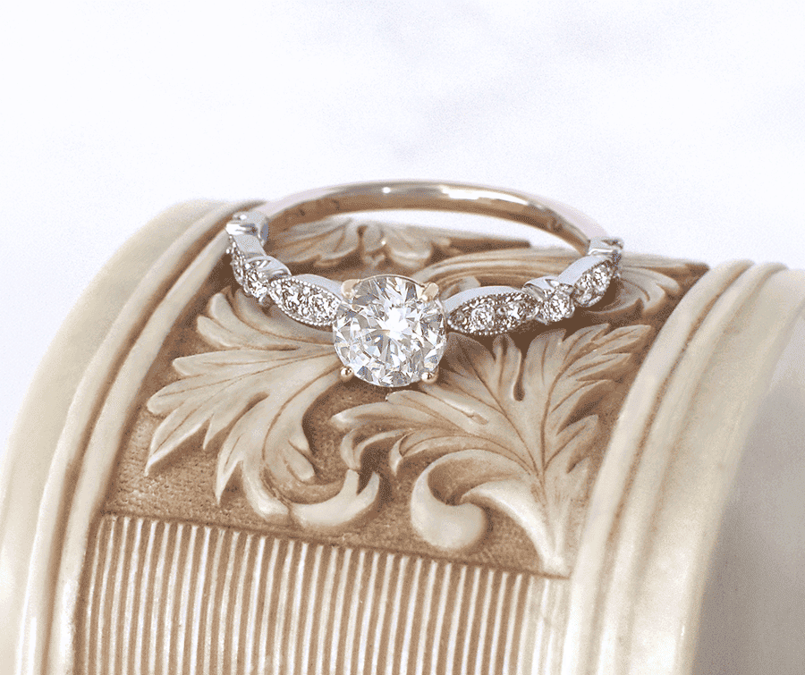 Vintage-inspired round brilliant diamond engagement ring with milgrain shoulder detailing resting on an ornate carved pedestal at Mark Solomon Jewellers.