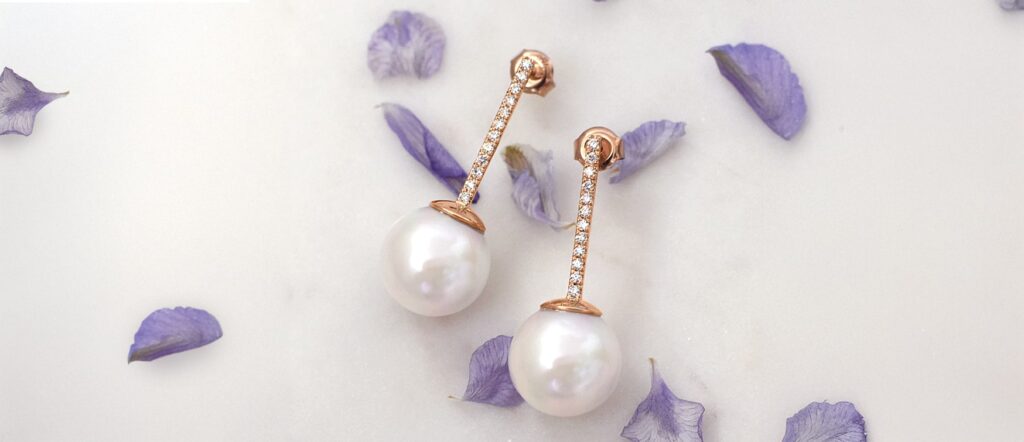 Rose Gold and Pearl Drop Earrings – Mark Solomon Jewellers - Mark Solomon Jewellers Rose Gold Drop Earrings with White Pearls and Diamond Accents on a White Surface with Purple Petals