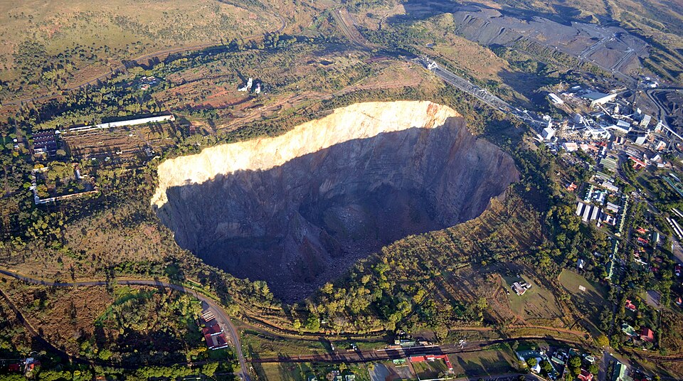 Aerial View of a Massive Circular Sinkhole or Crater in a Hillside, with Trees, Roads, and Buildings Around It.