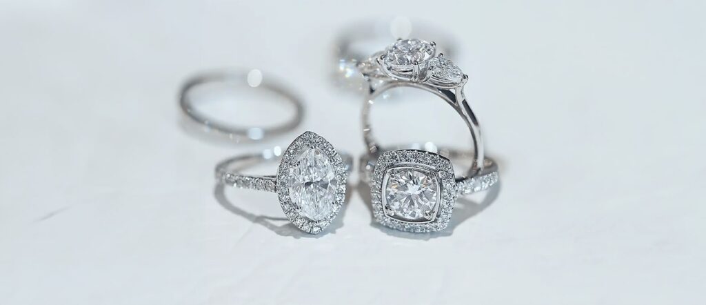 White Gold Diamond Engagement Rings – Trilogy and Halo Settings - Mark Solomon Jewellers Three Diamond Engagement Rings on a White Background – a White Gold Trilogy Ring with Round and Pear-shaped Diamonds, a Halo Ring with a Marquise-cut Diamond, and a Halo Ring with a Round Diamond and Cushion-shaped Halo.