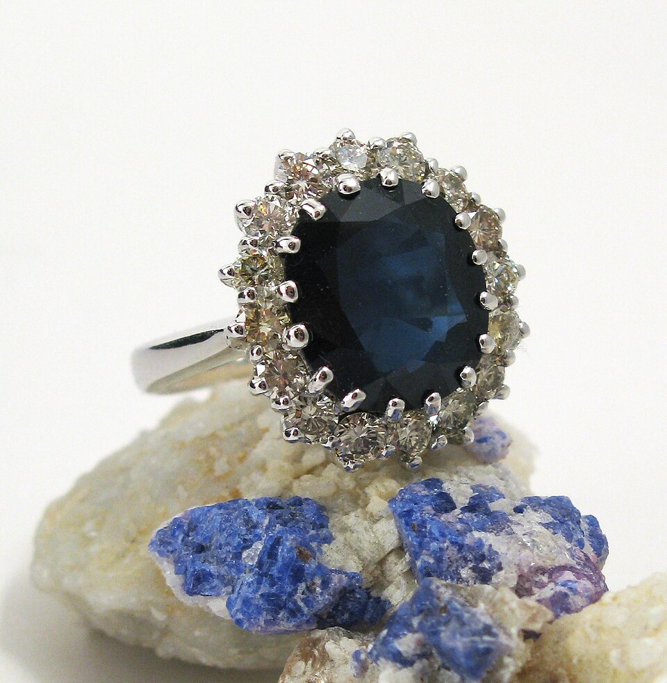 Silver Ring with a Large Oval Deep-blue Center Stone Surrounded by a Halo of Small Diamonds on a Rocky Display Surface, with Blue Crystal Accents.