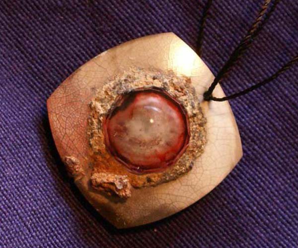 Beige Square Pendant Featuring a Glossy Red Cabochon Stone Surrounded by a Rough, Textured Ring on Purple Fabric Background.