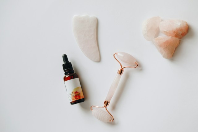 Rose Quartz Facial Roller and Gua Sha Stones with a Serum Bottle on a White Background.