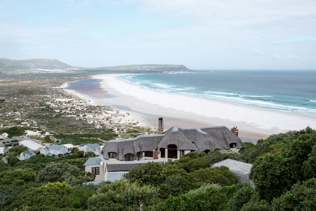 Coastal View of a Sandy Beach and Blue Ocean with a Thatched-roof House in the Foreground Among Green Shrubbery and Dunes