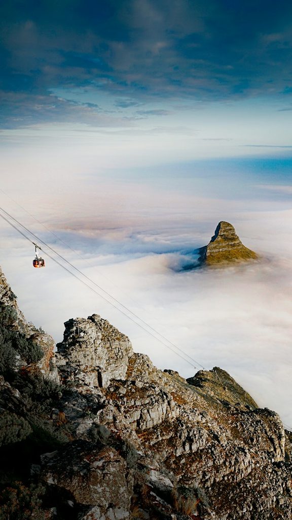 Cable Car Crossing Above Jagged Mountain Rocks with a Sea of Clouds and a Distant Peak in the Distance.
