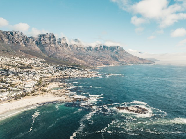John-o-nolan-rqrv0bbzmlg-unsplash - Mark Solomon Jewellers Aerial View of a Seaside Town Along a Rocky Coast with Mountains in the Background Under a Blue Sky.