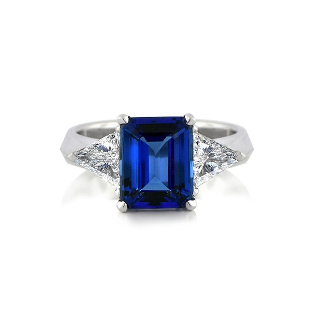 a Classic Three-stone Ring Featuring a Central Emerald-cut Deep Blue Tanzanite Flanked by Two Brilliant Triangle or Trillion-cut White Diamonds, Set in a Polished White Gold or Platinum Band.