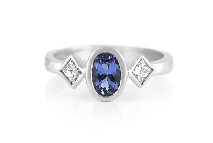 a Modern Three-stone Ring Featuring an Oval-cut Deep Blue Tanzanite Bezel-set in the Center, Flanked by Two Princess-cut White Diamonds Set in a Polished White Gold or Platinum Band.