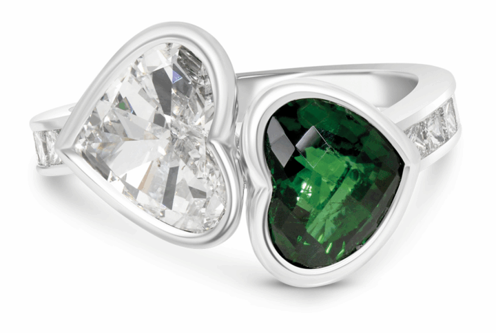 a Unique "toi Et Moi" Ring Featuring Two Heart-shaped Gemstones, a Clear Diamond and a Deep Green Gemstone (likely Emerald or Tourmaline), Bezel-set Side-by-side on a White Gold or Platinum Band with Channel-set Accent Stones.