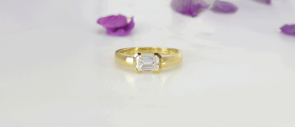 Mark Solomon Jewellers – Understanding Engagement Ring Prices in Cape Town - Mark Solomon Jewellers Yellow Gold Engagement Ring with an Emerald-cut Diamond, Set Against a Soft White Background with Scattered Purple Petals.