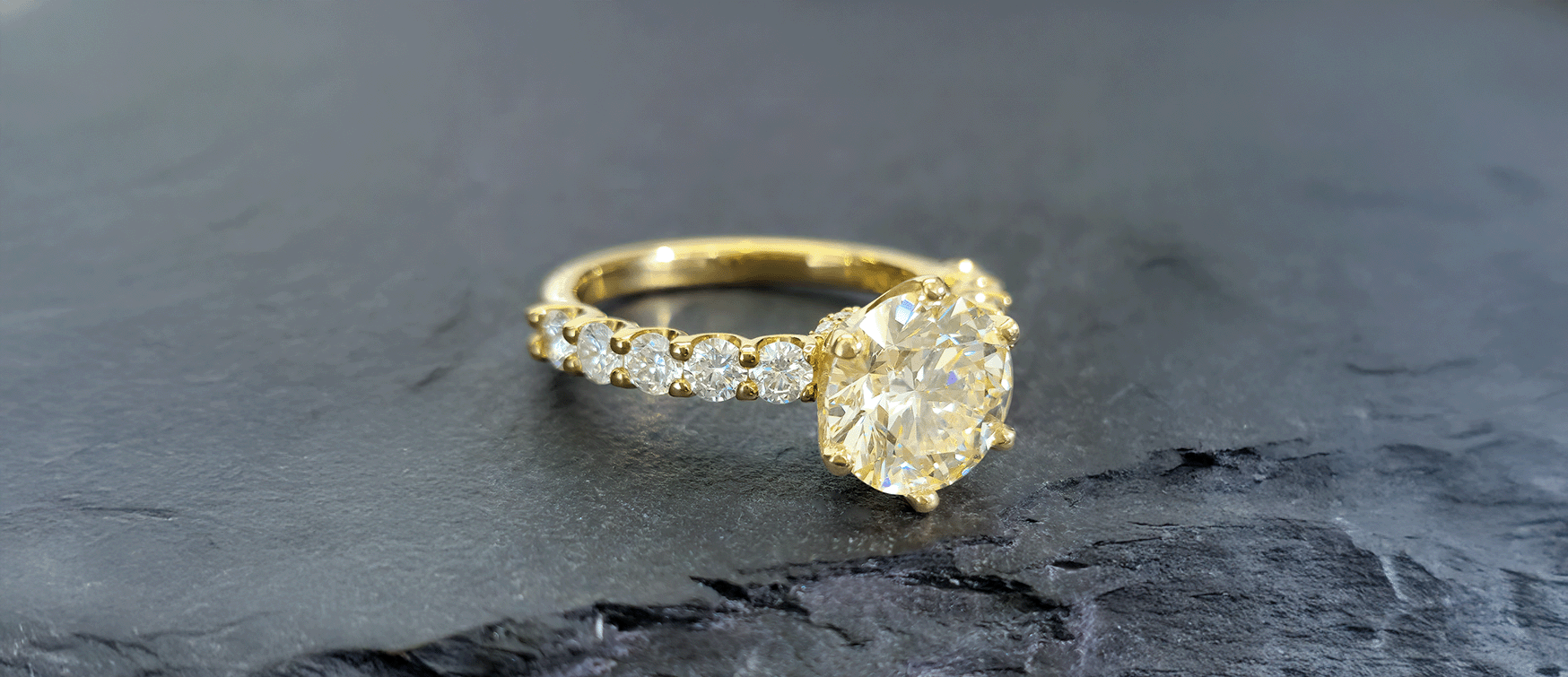 Oval-cut Fancy Yellow Diamond Engagement Ring Set in Yellow Gold with a Diamond Pavé Band, Photographed on a Dark Stone Surface.