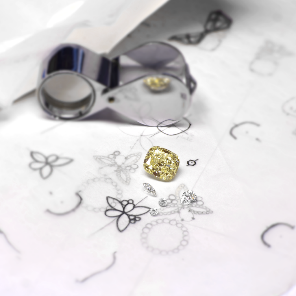 Yellow Cushion-cut Diamond and Small White Diamonds Resting on Hand-drawn Jewellery Sketches, Viewed Through a Metal Jeweller’s Loupe.