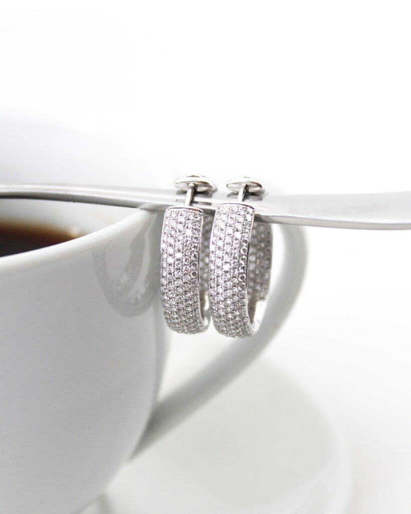 Broad_pave_huggie_earrings_daily_comfort - Mark Solomon Jewellers Diamond-studded Silver Hoop Earrings Resting on the Rim of a White Teacup, Close-up View.